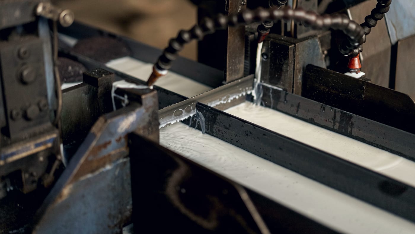An industrial bandsaw cutting through a steel section, coolant running over the blade.