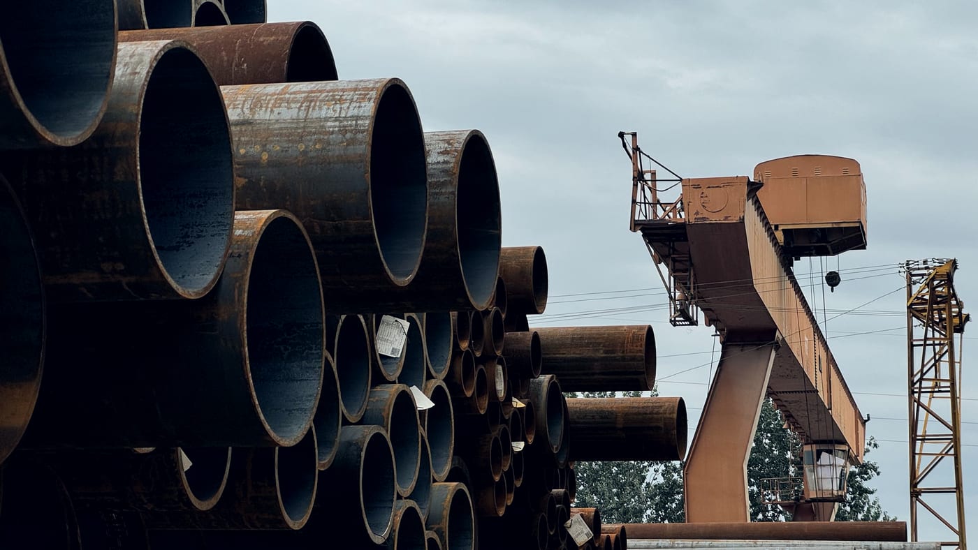 Stacked large-diameter steel pipe in an outdoor stockyard, with a gantry crane behind.